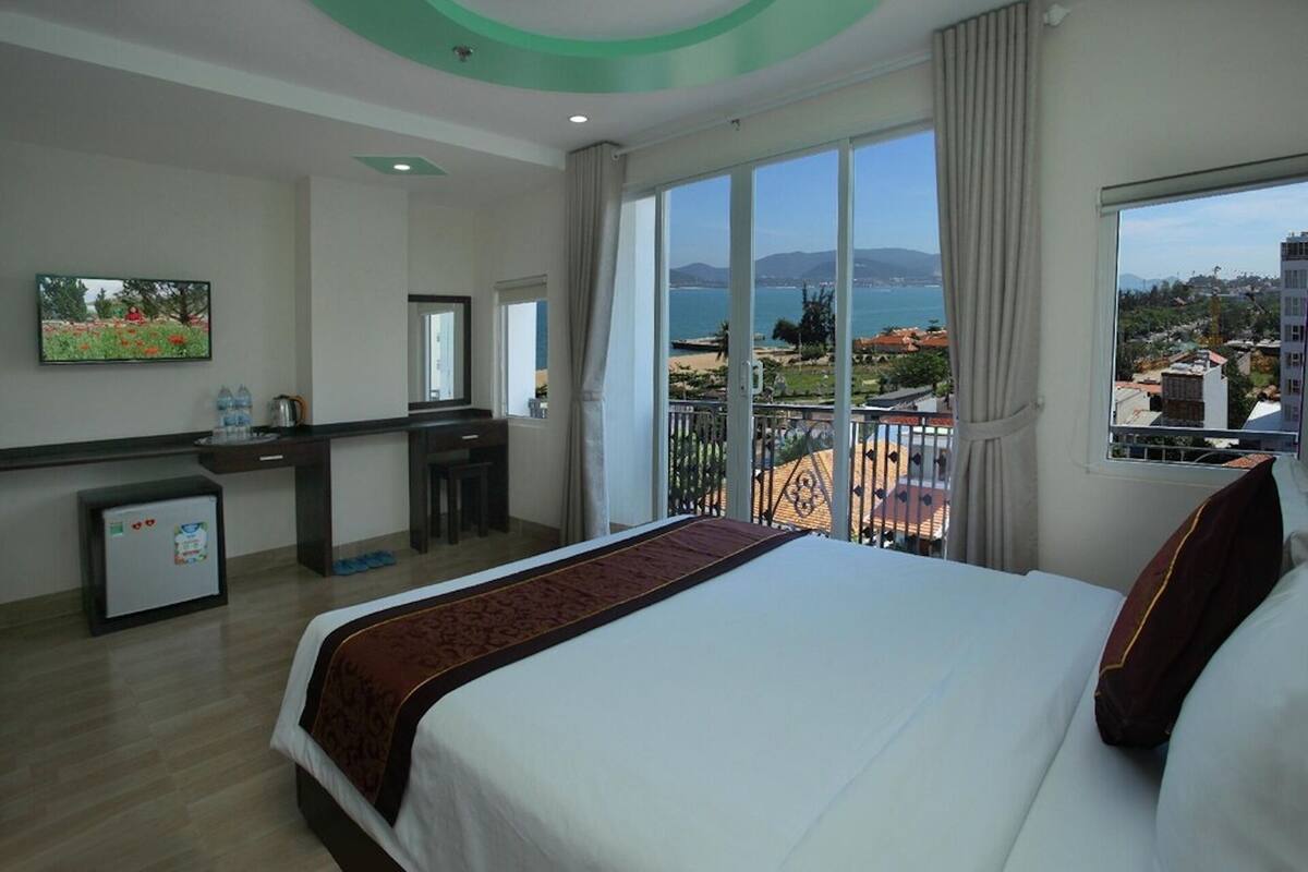 Une chambre d’hôtel équipée d’un lit, d’un bureau, d’un petit réfrigérateur et d’un balcon avec vue sur la ville.