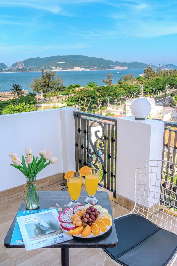 A balcony with a table set for breakfast, featuring a plate of fruit, a vase of flowers, and a magazine. The view includes a beach and hills in the distance. - Melyna Hotel Nha Trang Beach (Nha Trang)