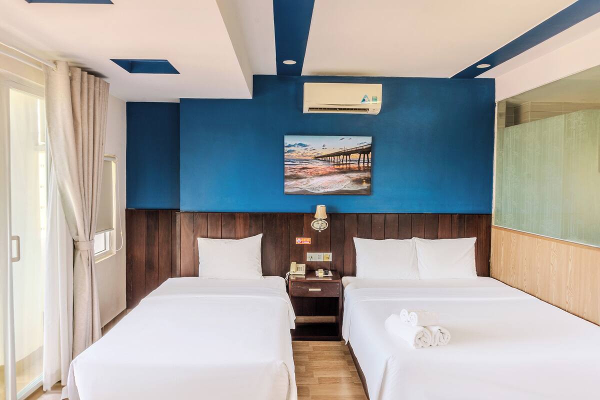 Une chambre d’hôtel avec deux lits, une tête de lit en bois, un climatiseur fixé au mur, un tableau représentant une jetée et une table de chevet avec une lampe.