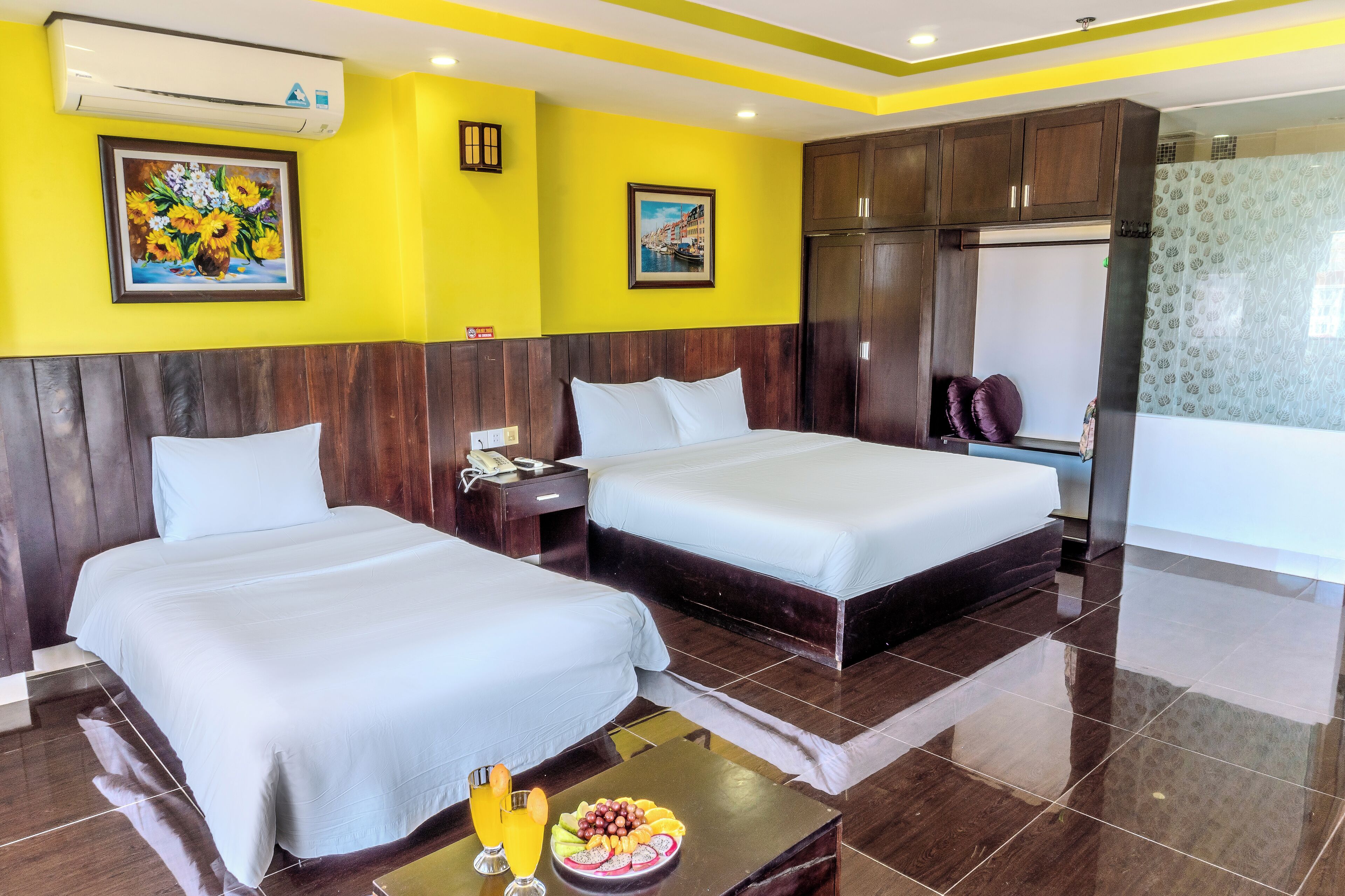 A hotel room with two beds, a wooden headboard, a small table with fruit, and a painting on the wall.