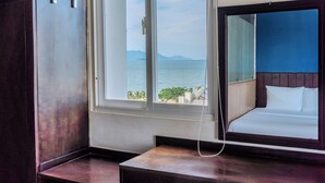 A hotel room with a large window offering a view of the sea and mountains, a bed with white linens, and a wooden nightstand. - Melyna Hotel Nha Trang Beach (Nha Trang)