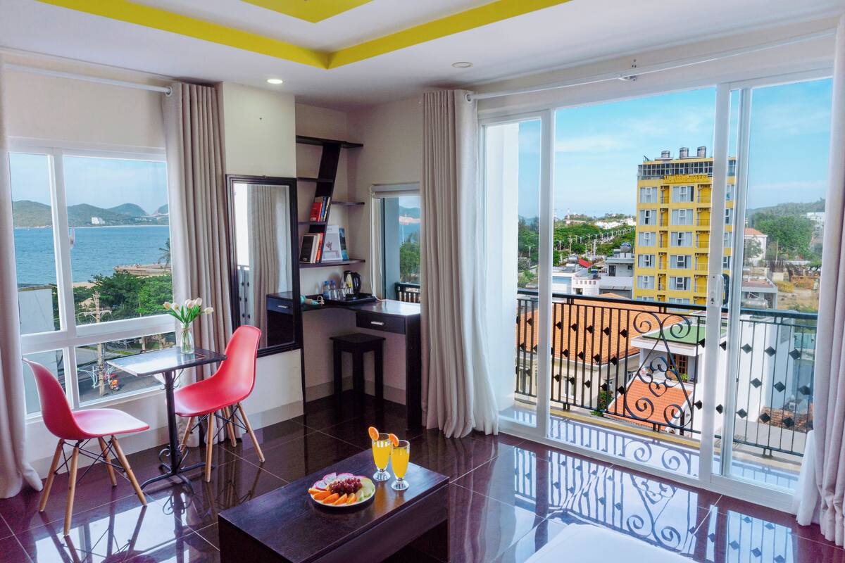 Une chambre d’hôtel moderne avec balcon, un coin repas avec des chaises rouges, une petite table avec une assiette de fruits et des boissons, et une vue sur la mer et les bâtiments.