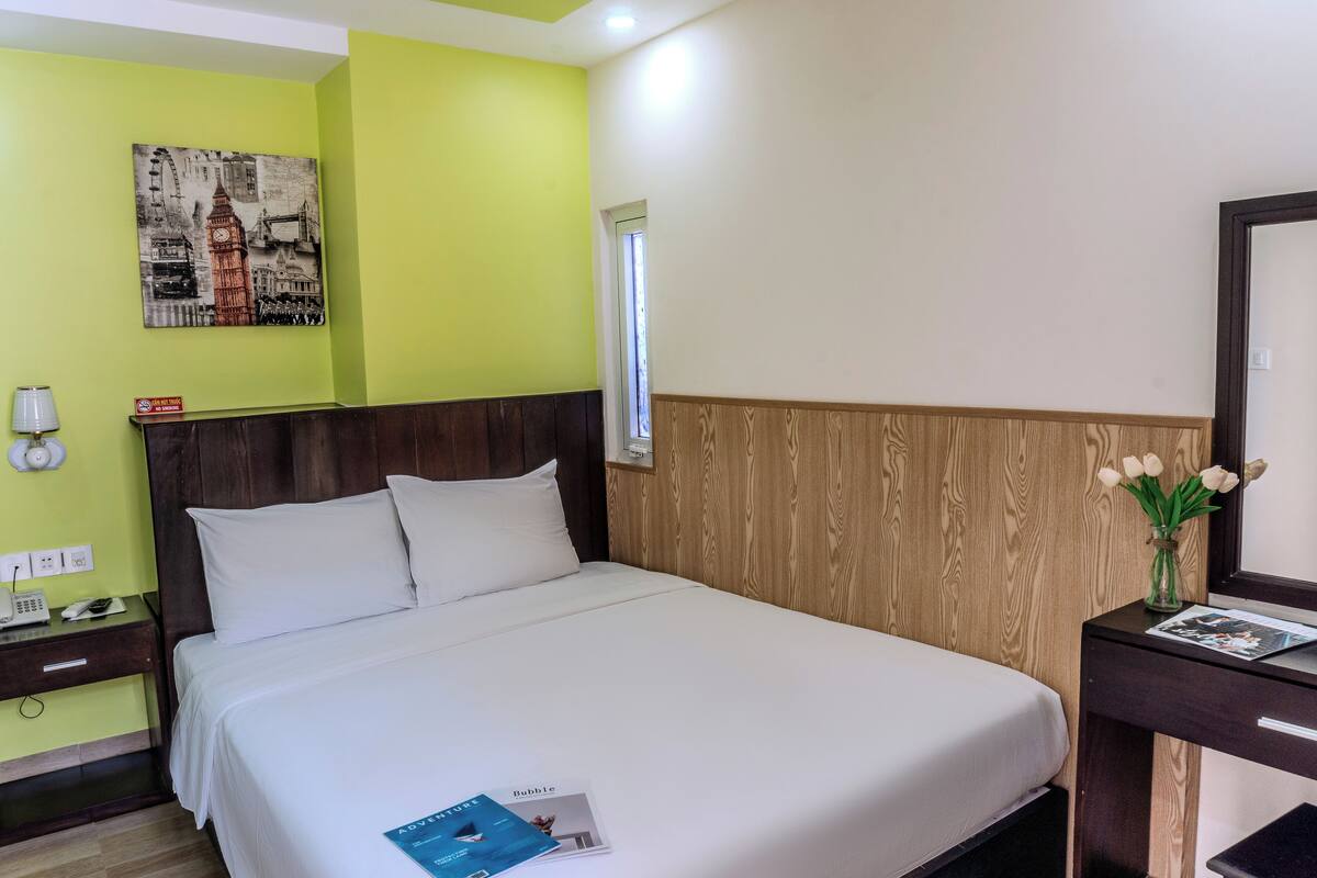 Une chambre d’hôtel avec un lit, une table de chevet, une tête de lit en bois, un miroir, un vase avec des fleurs et un tableau au mur.