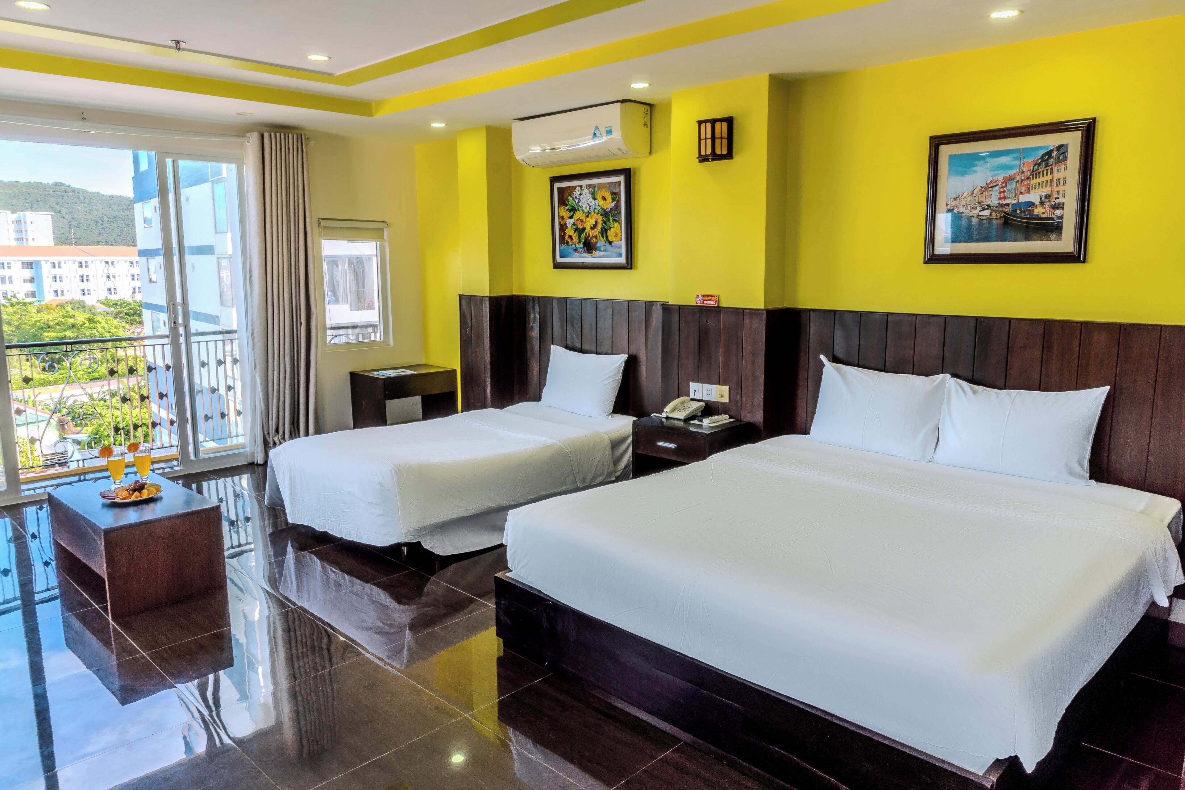 A hotel room with two beds, a small desk, a nightstand, and a balcony with a view of buildings and greenery.