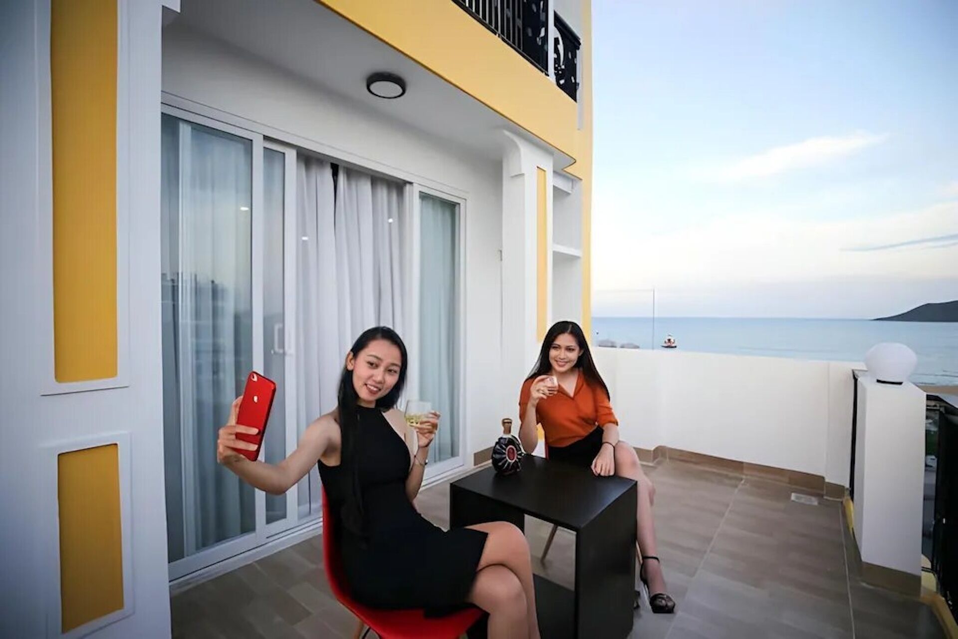 A balcony with two people taking a selfie, a table with a bottle, and a view of the sea.