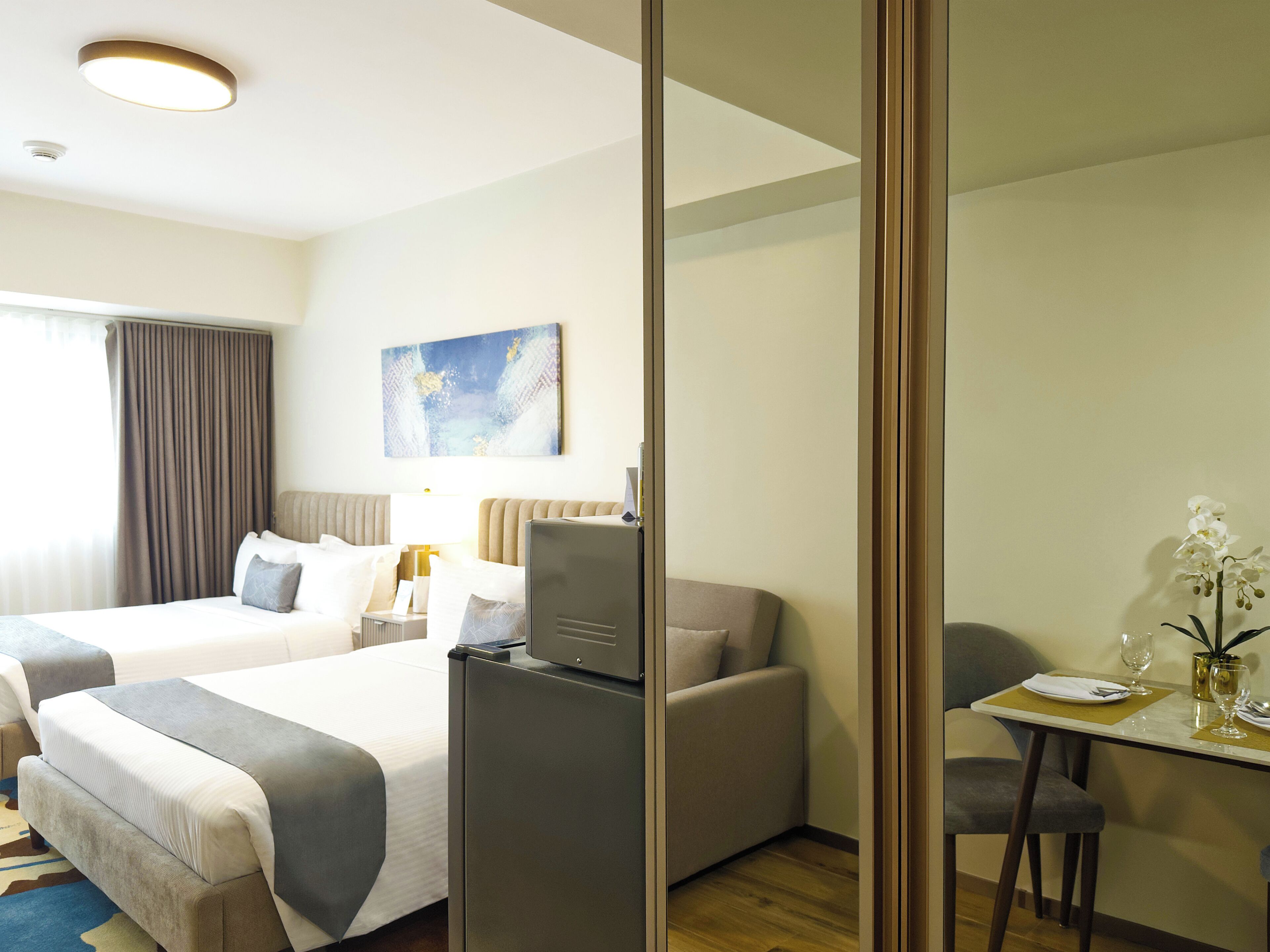 A hotel room with two beds, a desk, a chair, a refrigerator, and a painting on the wall.