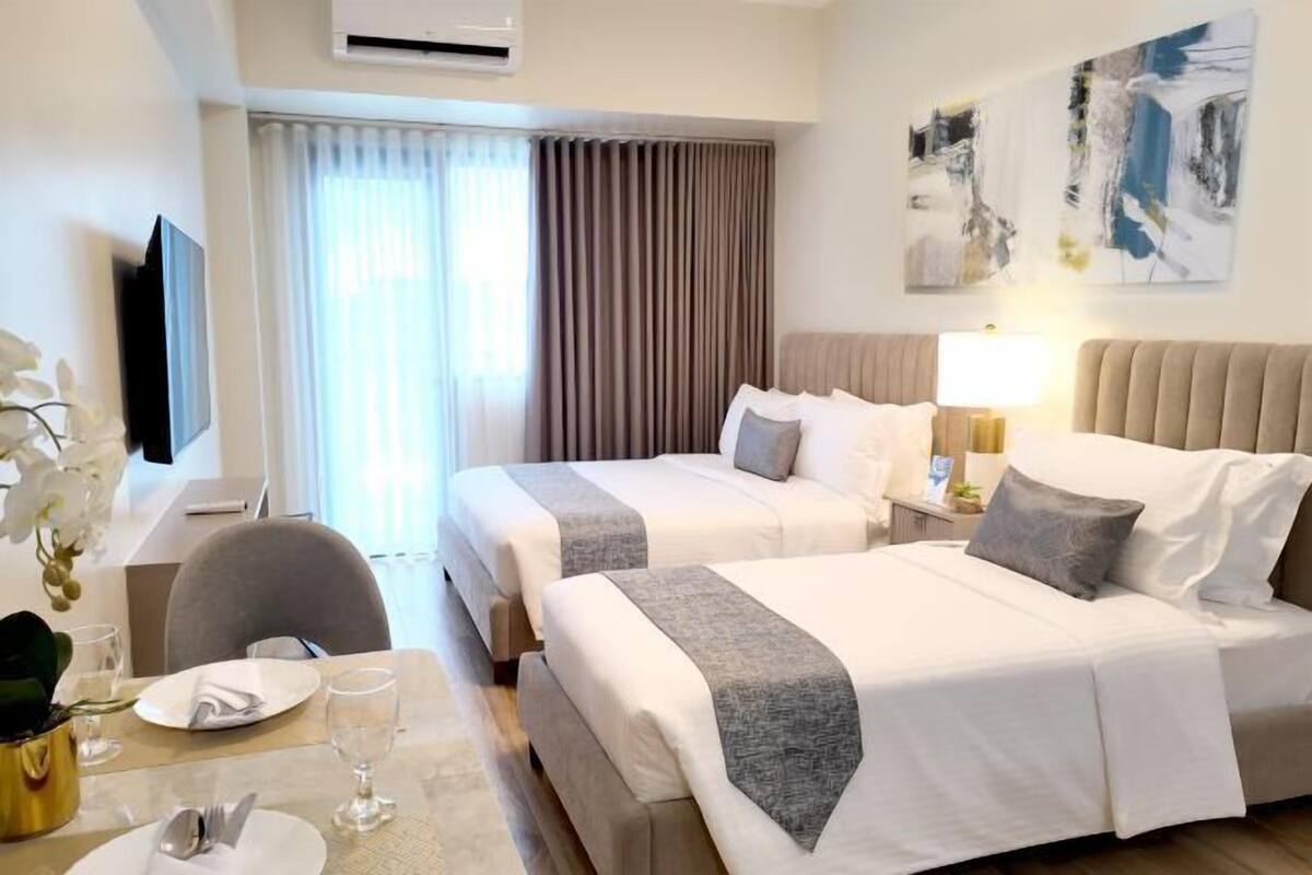 Habitación de hotel con dos camas, un televisor, una mesa de comedor con una silla y una ventana con cortinas.