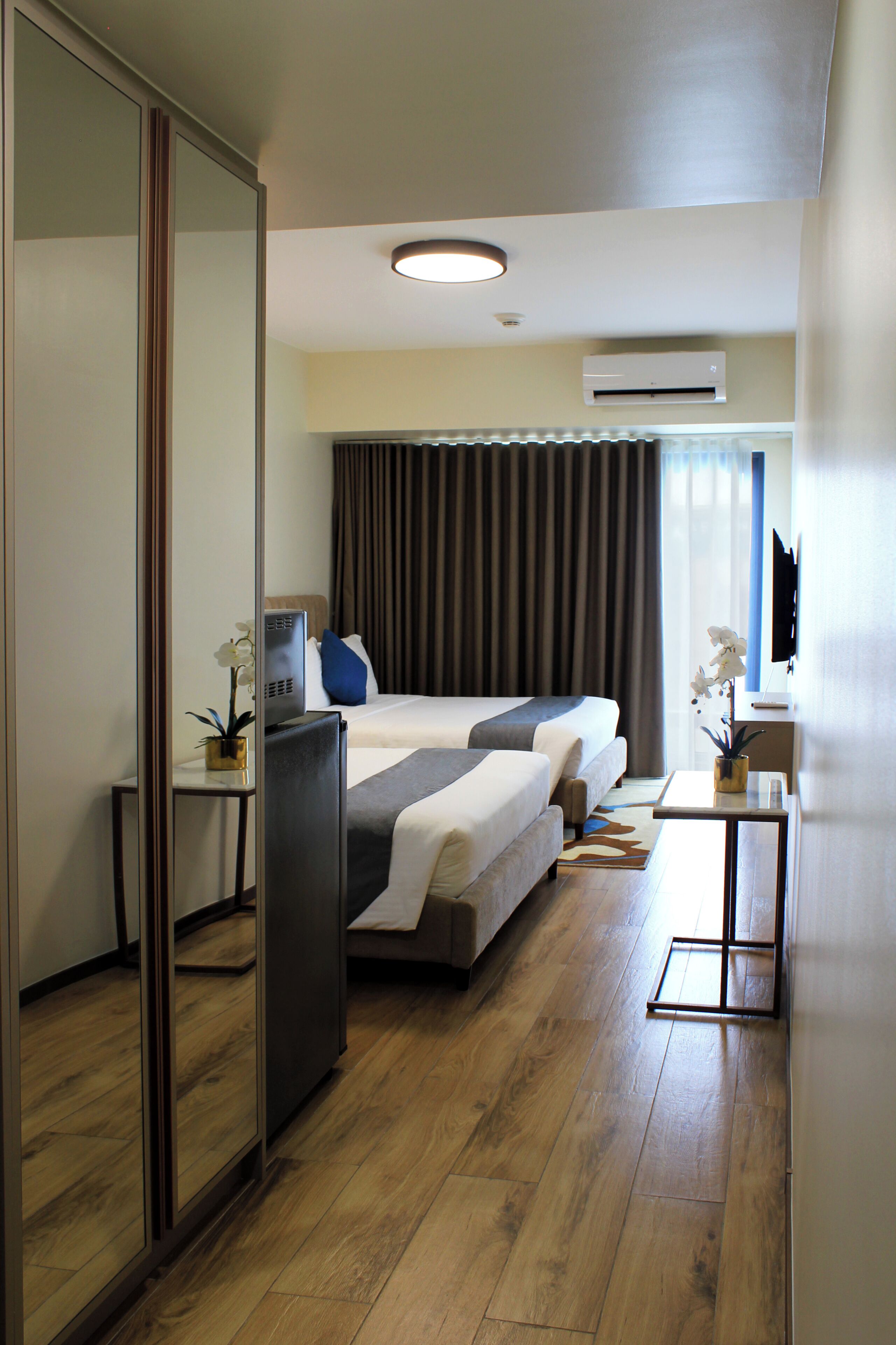 A hotel room with a bed, a desk, a mirror, and an air conditioning unit.