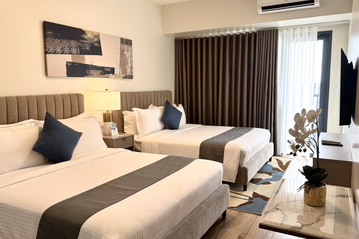 A hotel room with two beds, a desk, a TV, an air conditioner, and a large window with curtains.