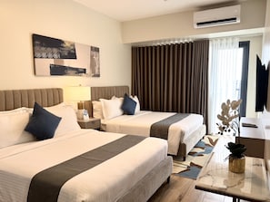 A hotel room with two beds, a desk, a TV, an air conditioner, and a large window with curtains.