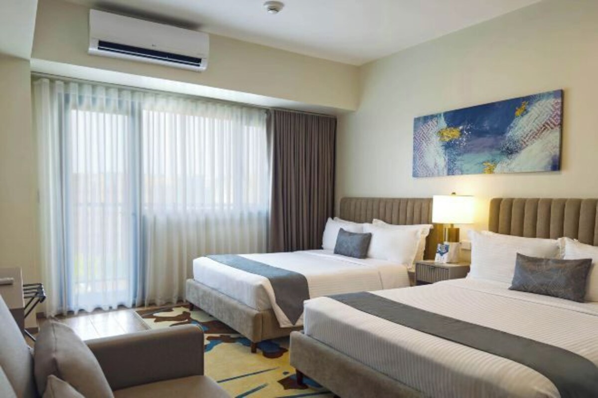 A hotel room with two beds, a desk, a chair, a window with curtains, and a painting on the wall.