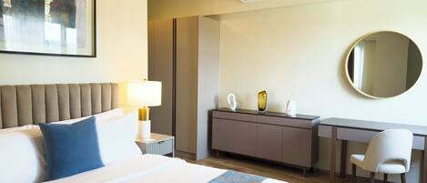 A hotel room with a bed, bedside table, lamp, desk, chair, and a round mirror.
