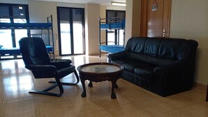 Una habitación con un sofá de cuero negro, un sillón de cuero negro y una mesa de centro de madera. Se ve una cama nido con ropa de cama azul al fondo.