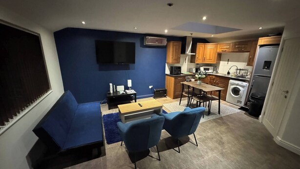 A compact living space with a blue accent wall, a flat-screen TV, a dining area with a table and chairs, a sofa, and a kitchen with wooden cabinets and appliances. - Tranquil Cambridge Getaway (Cambridge)