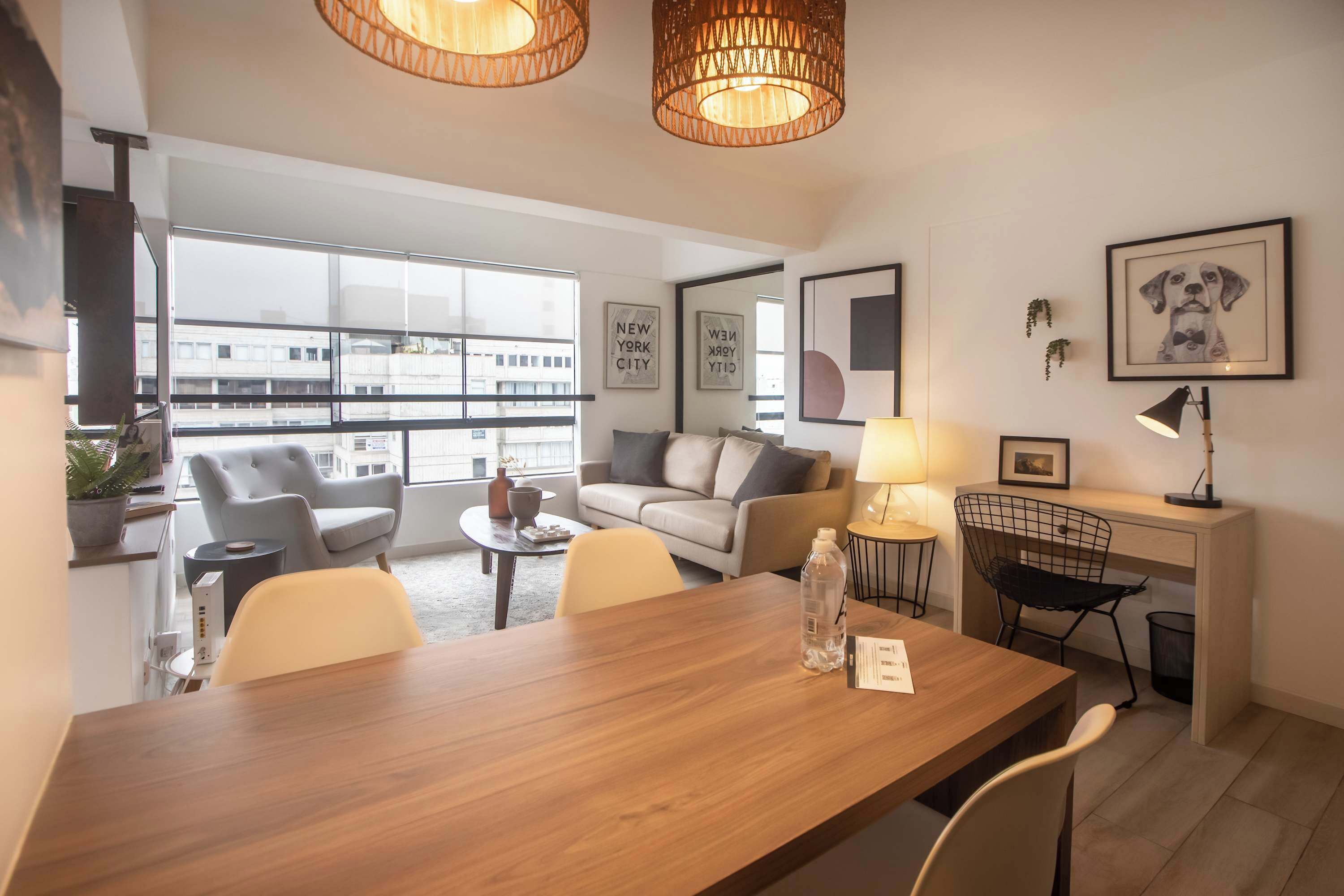 A modern living room with a wooden dining table, grey armchairs, a sofa, and a desk with a chair. There are framed pictures on the walls, a lamp, and a window with a view of buildings.