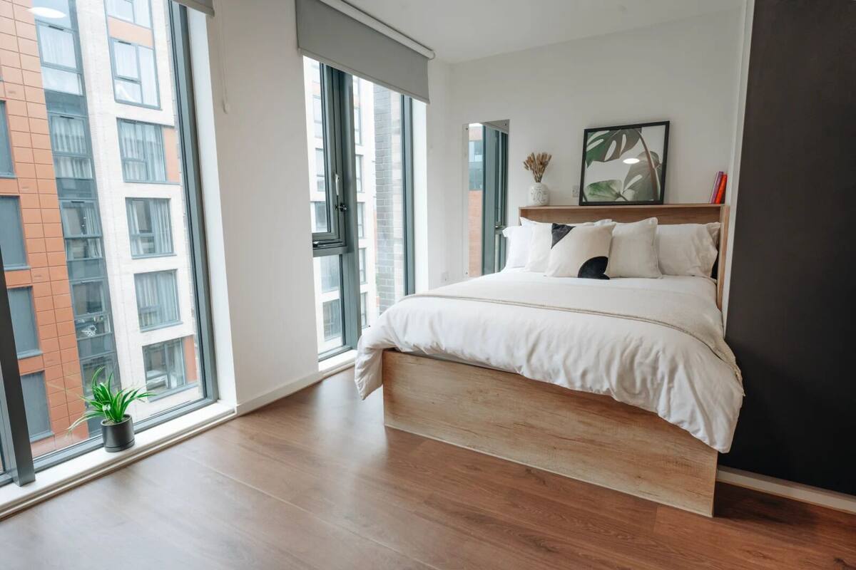 Ein modernes Schlafzimmer mit einem großen Bett, einem Holz-Kopfteil, einem kleinen Blumentopf und Blick auf eine Stadtlandschaft durch große Fenster.
