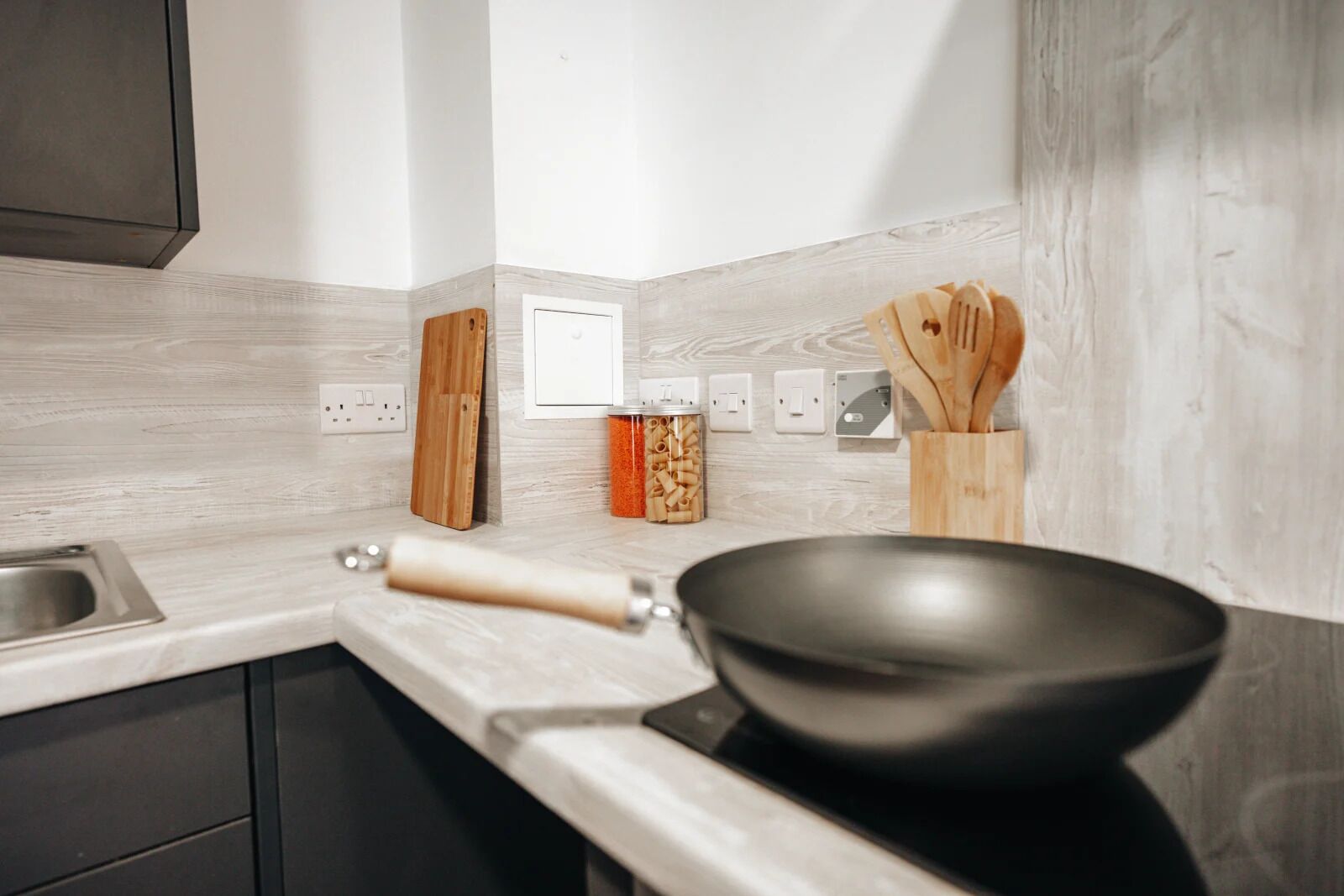 Een moderne keuken met een zwarte wok, houten keukengerei en een pastacontainer op het aanrecht.