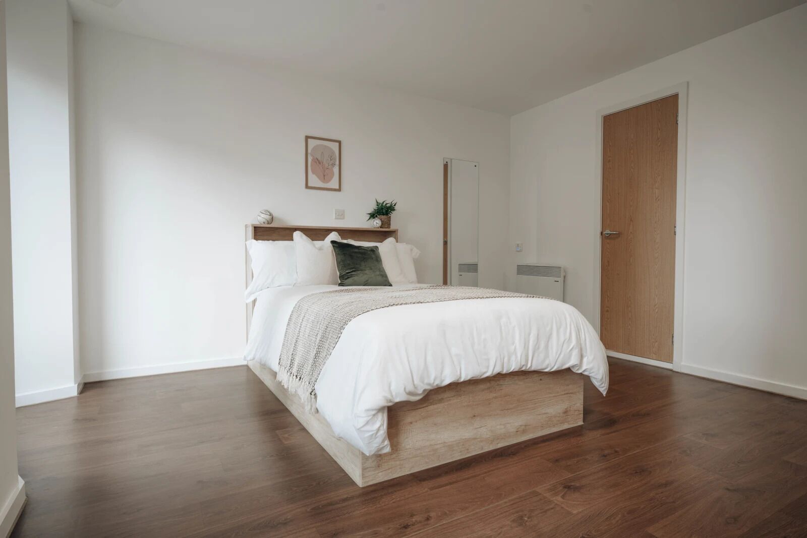 A bedroom with a wooden bed frame, white bedding, a small plant on the nightstand, and a wooden door.