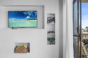 Un téléviseur à écran plat fixé à un mur blanc, une petite bibliothèque avec des livres, un panier en osier et une plante en pot.