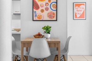 Un coin repas avec une table en bois, des chaises blanches, un saladier rempli de fruits, une plante en pot et deux œuvres d’art encadrées accrochées au mur.
