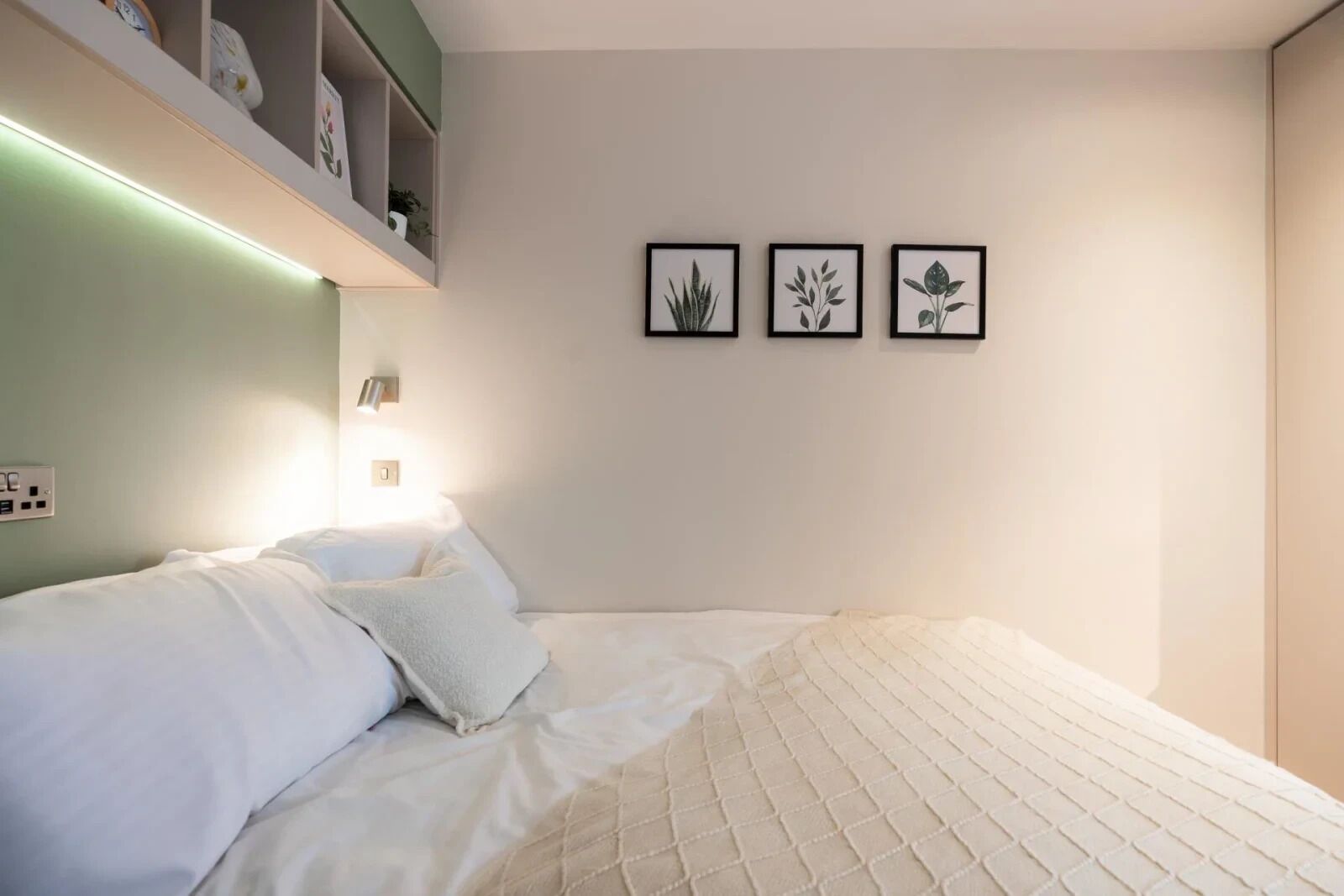 A neatly made bed with white linens and a quilted blanket, a bedside lamp, and a shelf with decorative items.