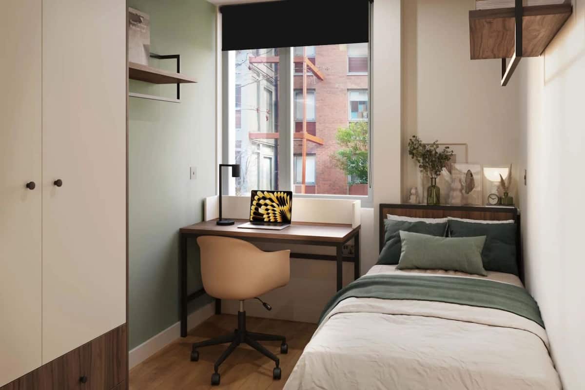 A bedroom with a bed, a desk, and a chair. There is a window with a view of buildings outside.