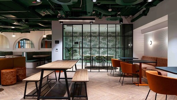 Un intérieur moderne avec des tables en bois et des chaises orange, un mur de verre offrant une vue dégagée et des éléments décoratifs verts au plafond.