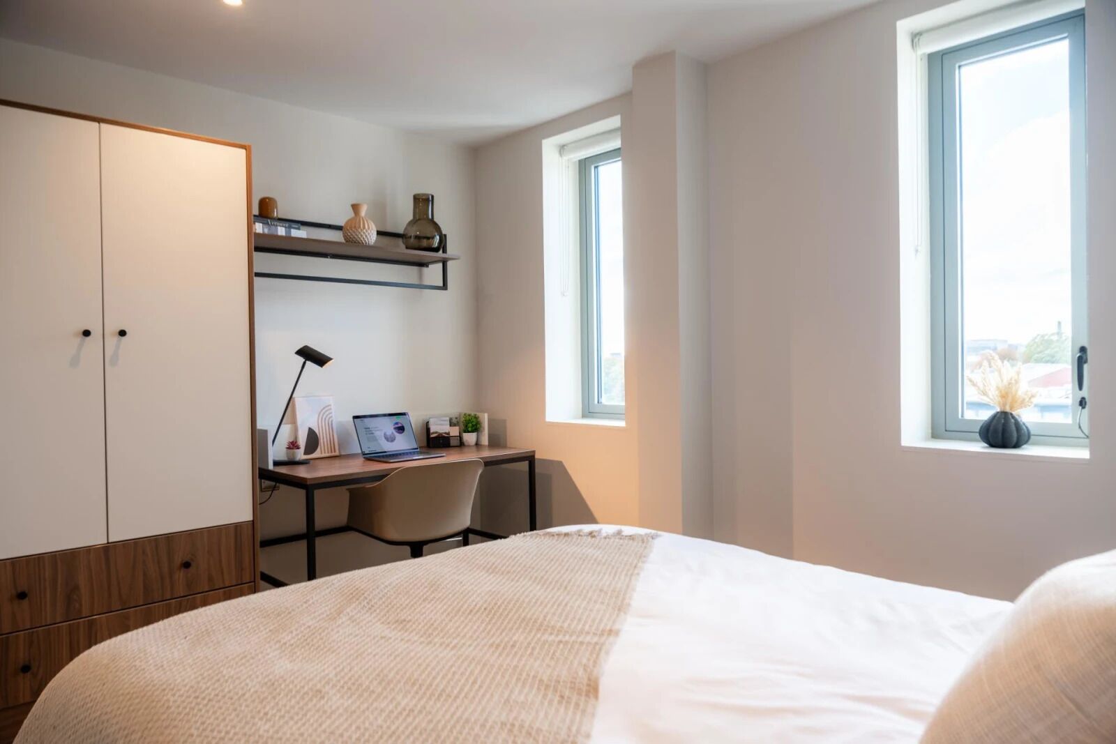 A bedroom with a bed, a desk with a laptop, a chair, a wardrobe, and a window.