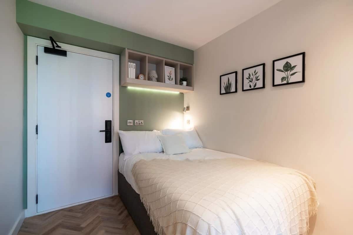 A bedroom with a bed, a white door, a shelf with books and a clock, and three framed pictures on the wall.
