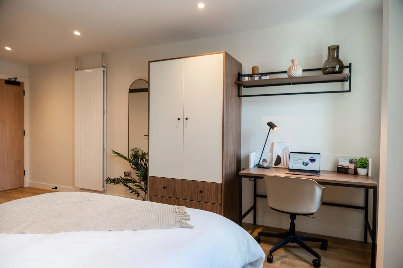 Een slaapkamer met een bed, een bureau met een laptop, een stoel, een kledingkast en een plank met decoratieve objecten.