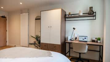 Een slaapkamer met een bed, een bureau met een laptop, een stoel, een kledingkast en een plank met decoratieve objecten.