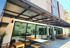 Un área de estar al aire libre con mesas y sillas, un porche cubierto y un edificio con ventanas amplias y techo de metal.