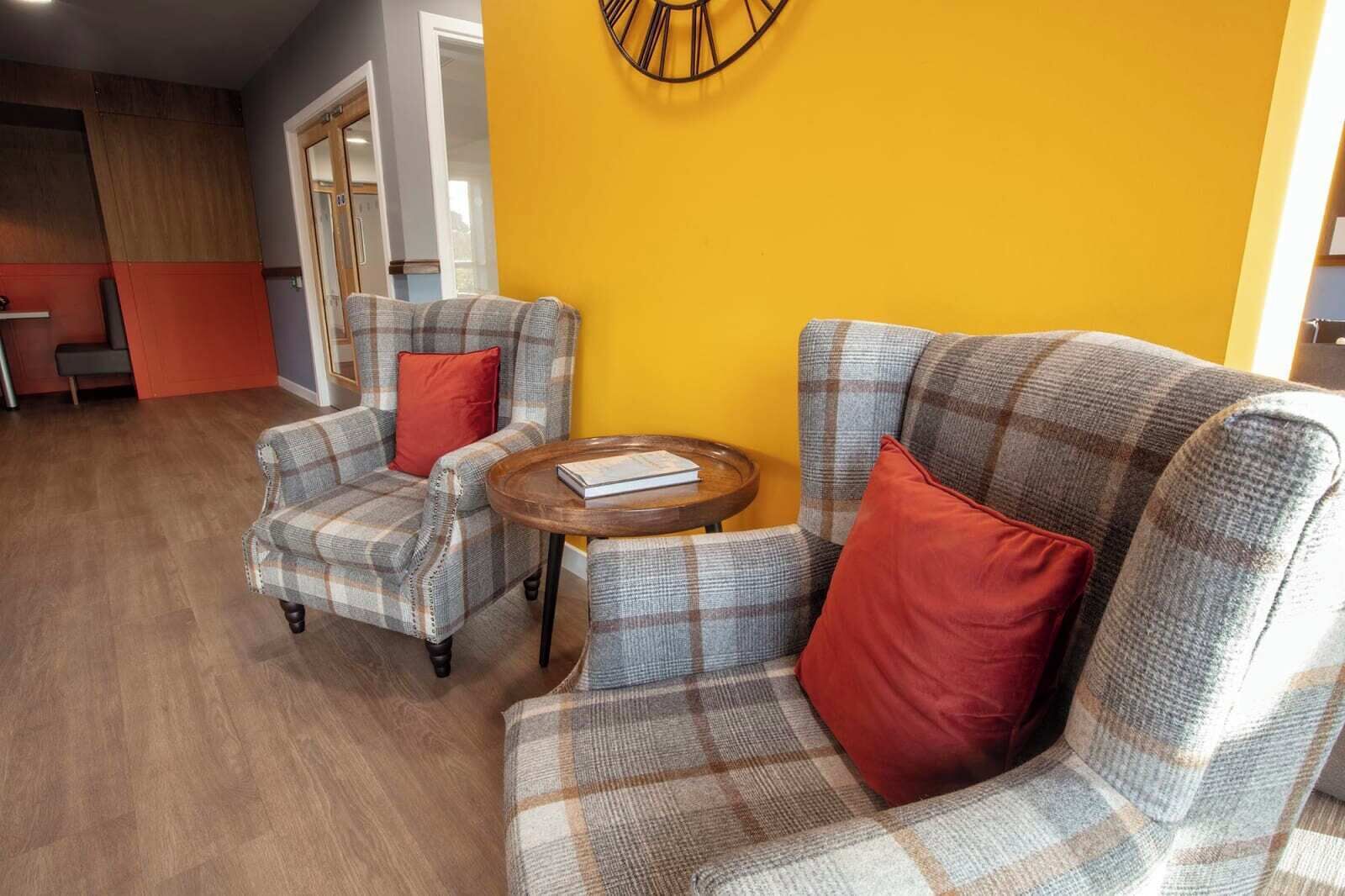 Een kamer met twee geruite fauteuils, een klein houten tafeltje en een gele accentmuur.