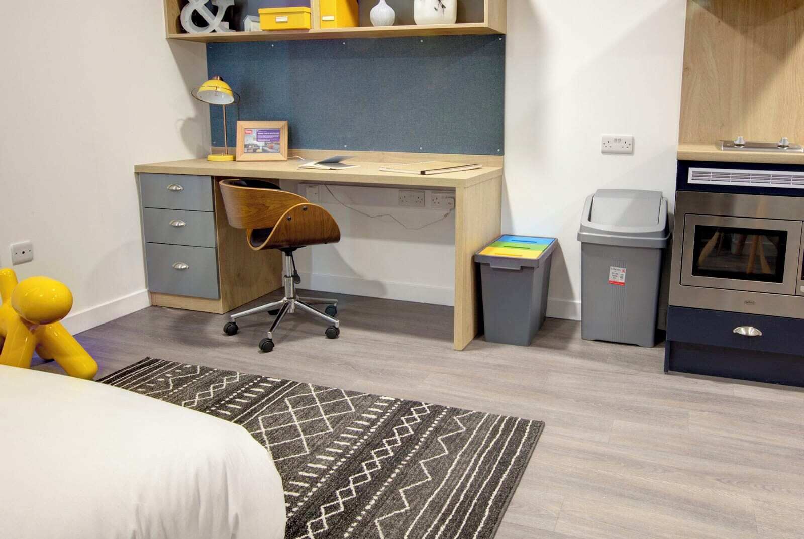 A compact bedroom with a bed, a desk with a chair, a small trash bin, and a kitchenette area.