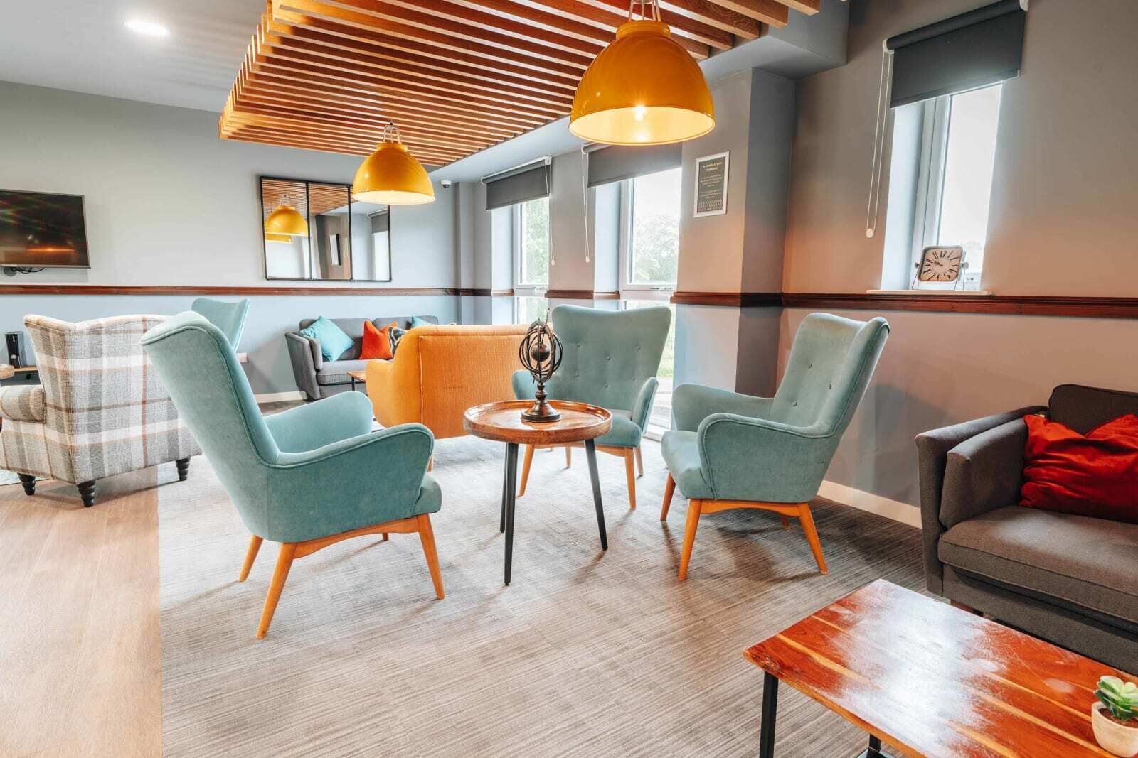 A modern lounge area with teal armchairs, a wooden coffee table, and a sofa with red cushions.