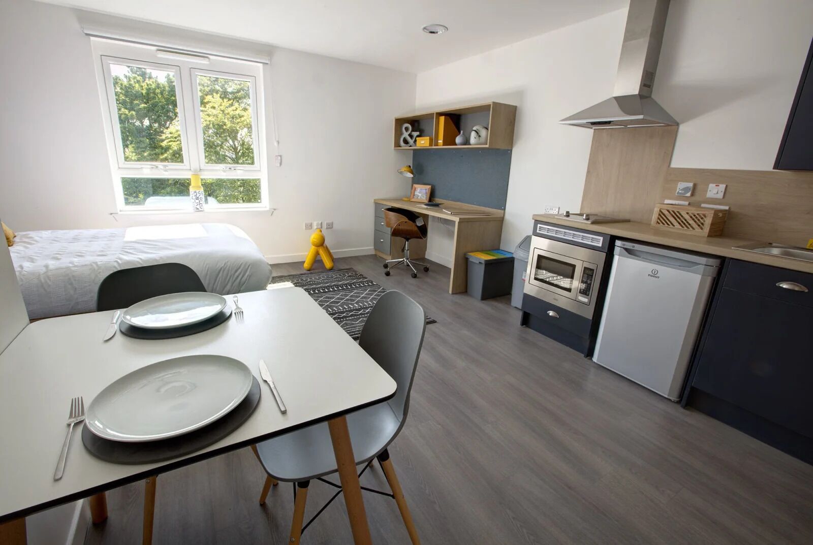 A compact, modern room with a bed, a dining table with chairs, a kitchenette with a microwave and oven, a desk with a chair, and a window with a view of trees.