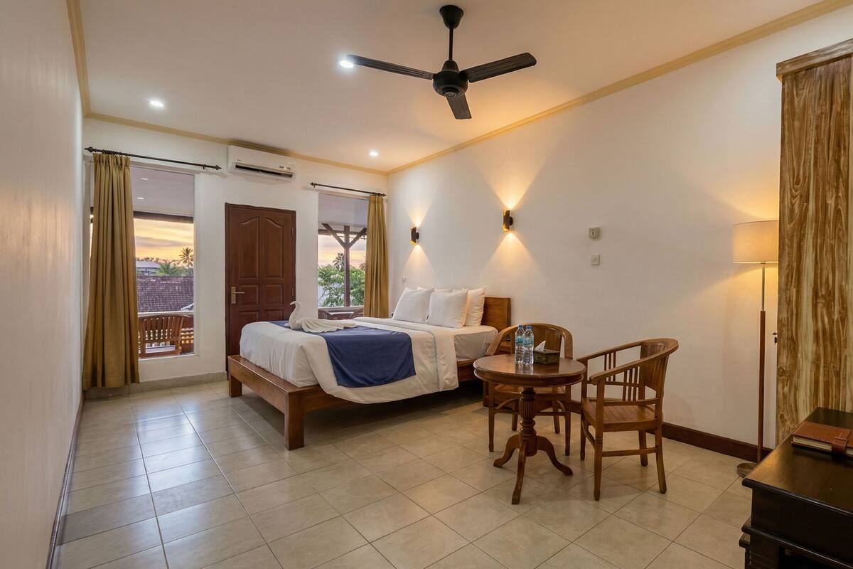 A hotel room with a bed, a table with chairs, a ceiling fan, and a view of the outdoors.