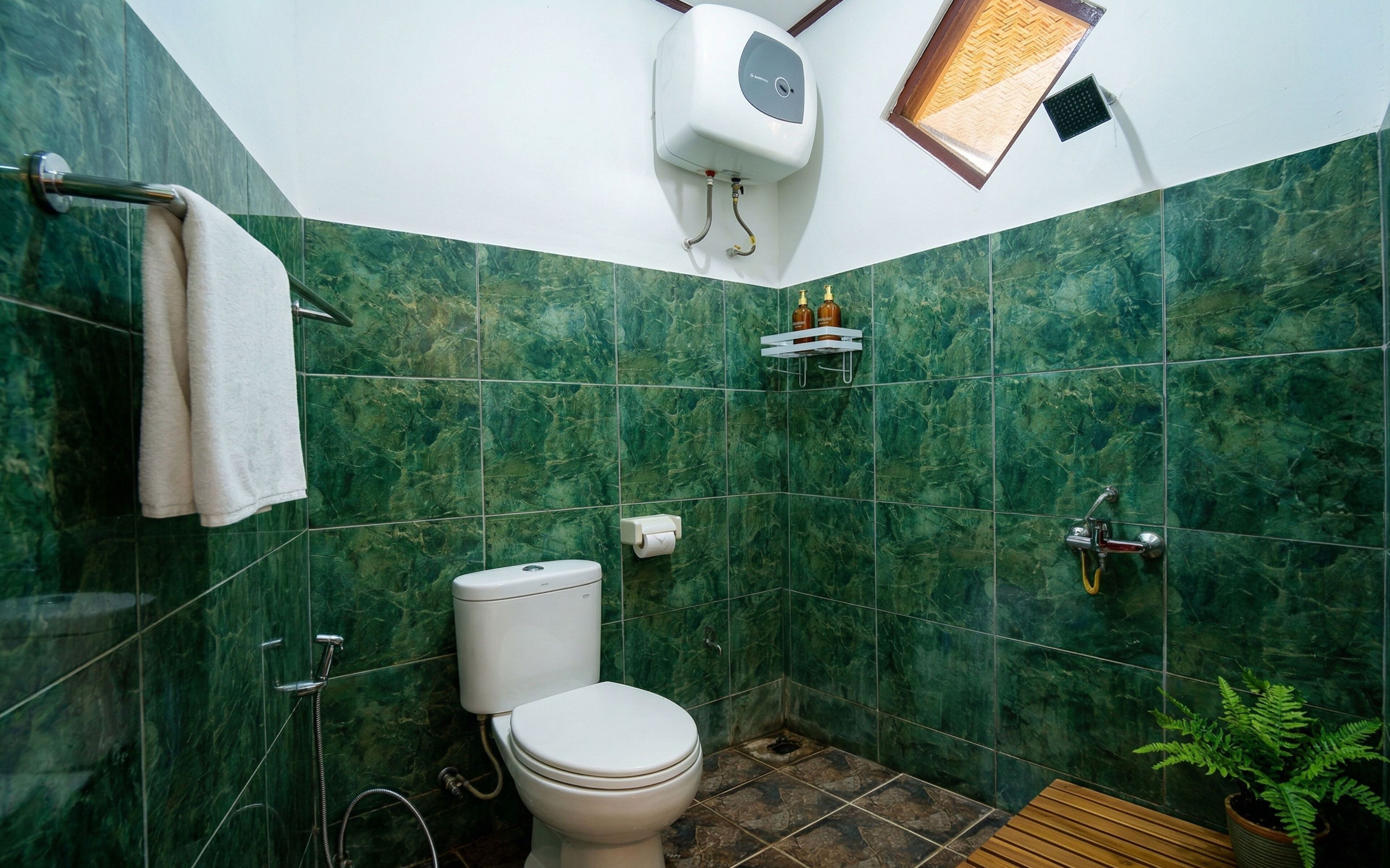 A bathroom with green marble tiles, a toilet, a shower, a towel rack, a soap dispenser, and a small plant.