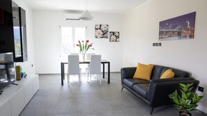 A modern living room with a black leather sofa, a dining table with chairs, a flat-screen TV, and a potted plant. - VivimiHome - Monti House (Monti)