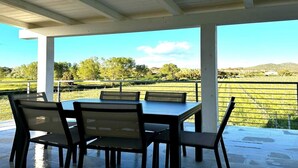 A covered outdoor seating area with a table and chairs, overlooking a green landscape with trees and hills. - VivimiHome - Monti House (Monti)