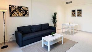 Una sala moderna con un sofá oscuro, una mesa de centro blanca y una lámpara de pie. Hay una mesita con una planta y dos cuadros en la pared.
