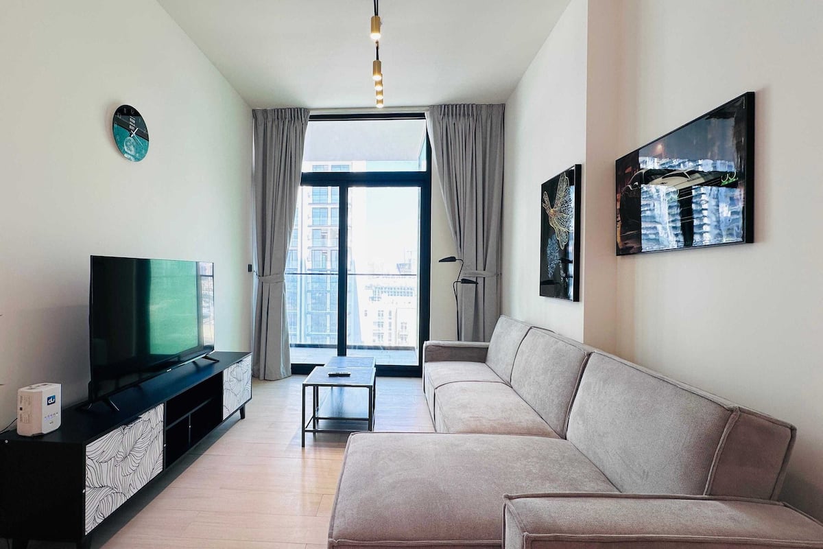 Un salon moderne avec un téléviseur à écran plat posé sur un meuble noir, un canapé beige, une petite table d’appoint et une grande fenêtre avec des rideaux.