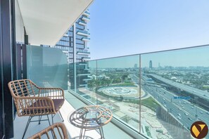 Een balkon met rieten stoelen en een glazen tafel, met uitzicht op een stadsgezicht met moderne gebouwen en een snelweg.