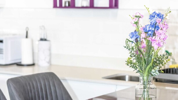 Une cuisine moderne avec une décoration murale violette, un vase transparent rempli de fleurs et un évier en acier inoxydable.