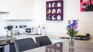 Une cuisine moderne avec un coin repas, des meubles blancs, un porte-épices violet et un vase rempli de fleurs posé sur le plan de travail.