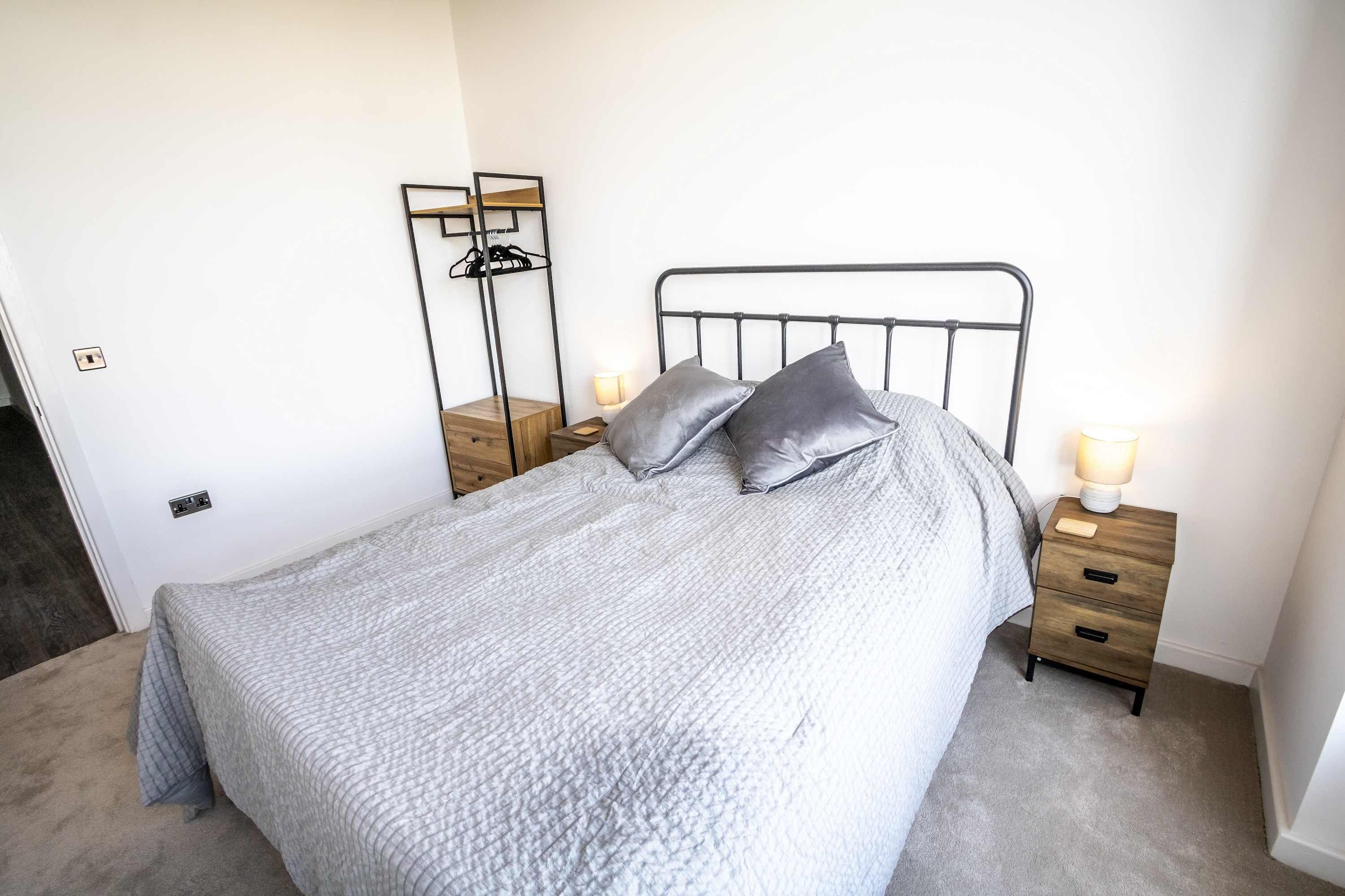 Un lit bien fait, recouvert d’une couette grise, deux tables de chevet avec des lampes, et une petite table en bois avec un miroir et des étagères.