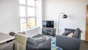Un salon moderne doté d’une grande fenêtre, d’un téléviseur à écran plat posé sur un meuble en bois, d’une lampe sur pied et de deux canapés gris agrémentés de coussins.