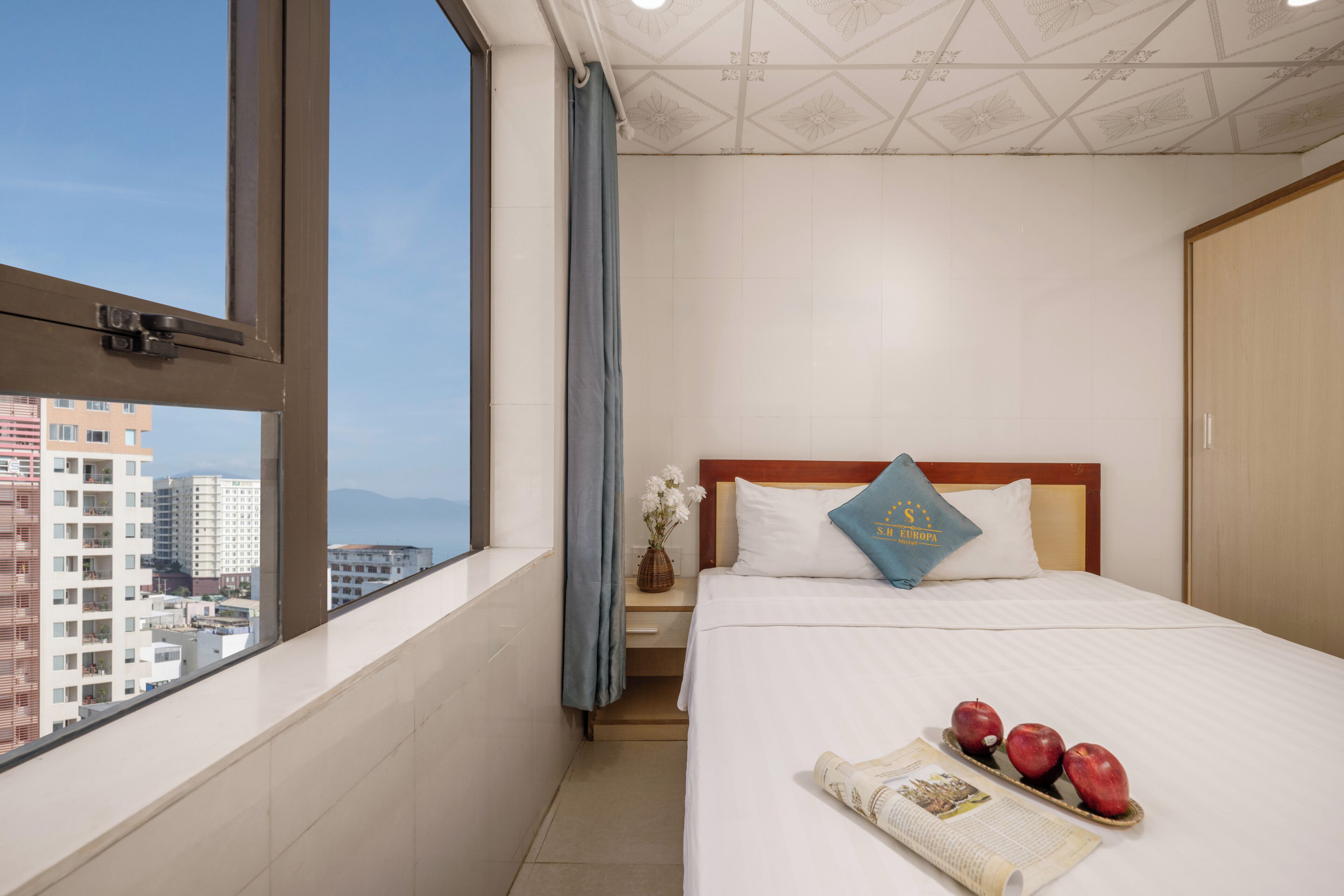 A hotel room with a bed, a nightstand, a window with a view of buildings, and a tray with apples on the bed.