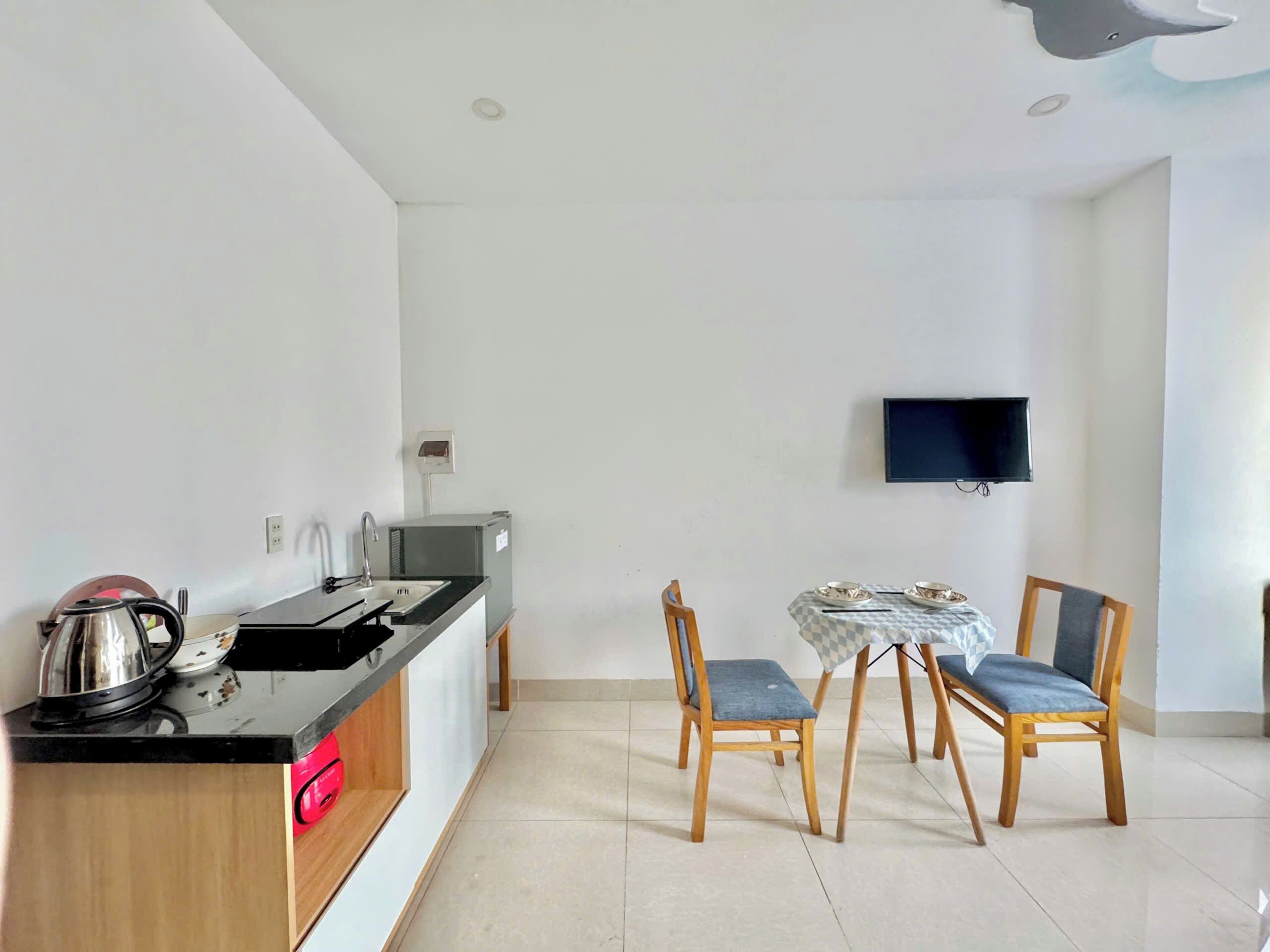 Una cocina compacta con un lavamanos, hervidor de agua y un refrigerador pequeño. Hay una mesa de comedor con sillas y un televisor de pantalla plana montado en la pared.