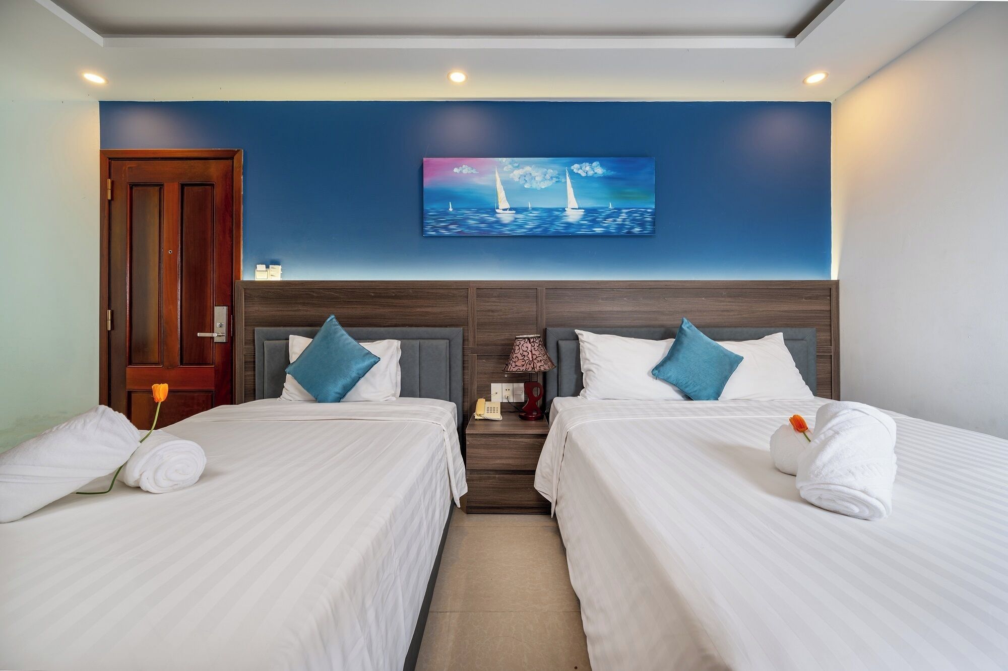 A hotel room with two beds, a painting of sailboats on the wall, a wooden door, and a nightstand with a lamp and phone.